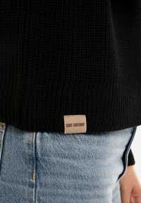 Zwarte gebreide trui met ribbelstructuur, voorzien van een klein leren label met de tekst "Knit Factory" aan de zoom, gecombineerd met lichtblauwe spijkerbroek.