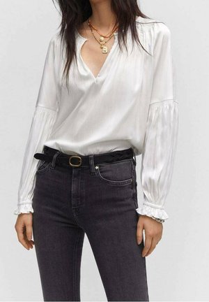 Kobieta nosząca białą jedwabną bluzkę z długim rękawem z marszczonymi mankietami, ciemne dżinsy z wysokim stanem, czarny pasek i warstwowe złote naszyjniki.