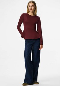 Sieviete, tērpusies piegulošā burgundijas krāsas garajā piedurknē, brīvās tumši zilās džinsās un zaļās kurpēs, stāv ar vienu roku kabatā uz balta fona.