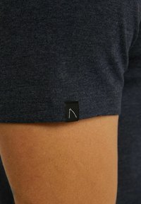 Tmavomodré tričko s krátkym rukávom a hladkou textúrou. Obsahuje malú čiernu nálepku s bielym grafickým detailom pri leme.