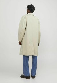 Ljusbeige oversized-kappa med krage, långa ärmar och en bakre ventspringa, ihop med blå raka jeans och svarta skor.
