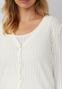 Witte geribde cardigan met een v-halsontwerp, voorzien van knopen aan de voorkant en een zachte textuur, gelaagd over een witte geribde top.