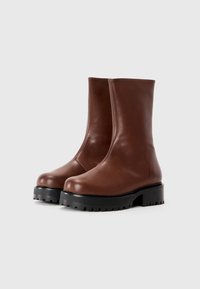 Par bok srednjedužih smešnih čizama od smešnog braon kože sa debelim crnim đonovima, zaobljenim vrhovima i glatkom površinom na jednostavnoj beloj pozadini.