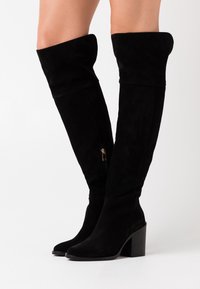 Botas de gamuza negra por encima de la rodilla con un dobladillo ancho, detalle de cremallera dorada y un tacón ancho negro. Textura suave y forma esbelta.