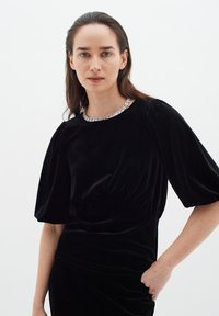 Abito in velluto nero con maniche a palloncino corte e colletto rotondo decorato con dettagli in argento. Tessuto liscio con una vita arricciata.