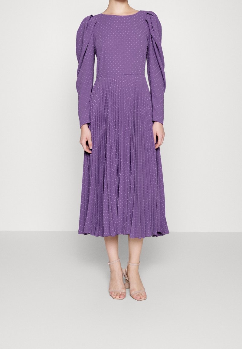 Femme portant une robe midi violette à manches longues avec épaules bouffantes, petit motif à pois et jupe plissée, associée à des talons à bride cheville couleur nude.