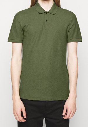 Person, die ein olivgrünes Poloshirt mit kurzen Ärmeln, Kragen und zwei Knöpfen trägt, kombiniert mit schwarzen Hosen vor einem einfarbigen Hintergrund.