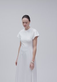 Femme aux cheveux foncés attachés porte une robe blanche simple à manches courtes et un bracelet argenté, debout devant un fond gris clair.