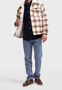 Beige en bruin geruite overhemdjas met een zachte voering, een zwarte button-up shirt eronder, blauwe jeans en bruine veterschoenen.
