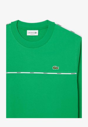 Camiseta de algodón verde con cuello redondo, franja blanca con "LACOSTE" y un logo de cocodrilo en el lado izquierdo del pecho. Diseño de ajuste clásico.