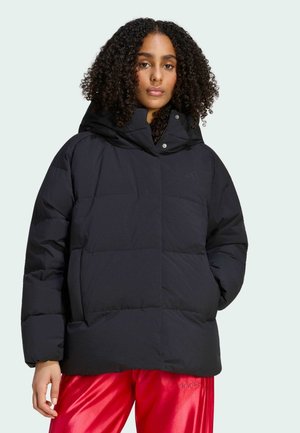 Femme aux cheveux bouclés portant une veste noire matelassée à capuche et un pantalon rouge brillant, debout devant un fond uni.