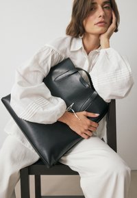 Borsa a tracolla nera in pelle con una texture liscia, dotata di manico superiore e dettaglio di chiusura. Il modello indossa una camicia bianca dal taglio ampio.