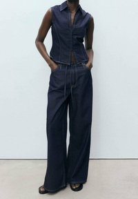 Jeansweste mit geradem Schnitt und Kragen, kombiniert mit weiten Jeanshosen. Dunkelblauer Stoff mit sichtbaren Nahtdetails.