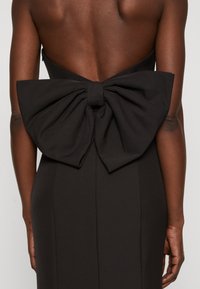 Schwarzes Kleid mit tiefem Ausschnitt auf der Rückseite, versehen mit einer großen Schleifendetail an der Taille. Glattes, mattes Material mit taillierter Passform, das die Sanduhrfigur betont.