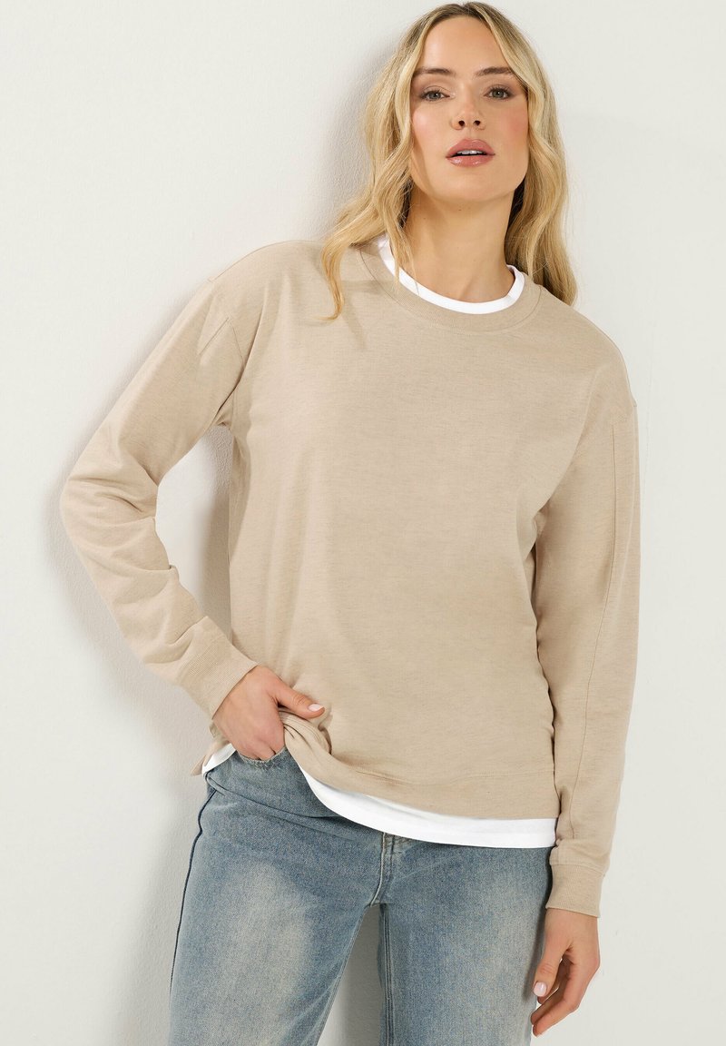 Blonde Frau mit welligem Haar trägt ein beiges langärmeliges Hemd über einem weißen Oberteil und hellblaue Jeans, steht vor einer schlichten weißen Wand.