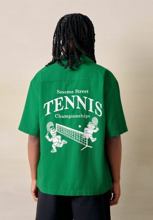 Person with long dreadlocks wears a green shirt with white "Sesame Street Tennis Championships" text and cartoon characters playing tennis.
