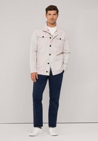 Giacca colletto grigio chiaro con quattro tasche frontali, bottoni neri e tessuto liscio, abbinata a pantaloni blu navy e sneakers bianche.