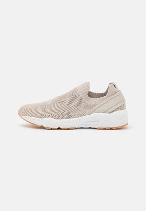 Beige slip-on kenkä, jossa on teksturoitu neulepäällinen, pehmeä kanta ja valkoinen pohja, jossa on kuminen pinta pidon parantamiseksi.