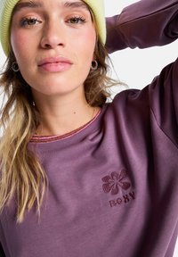 Sudadera morada con un acabado texturizado, que presenta un cuello a rayas rosas y el logo de Roxy bordado en el pecho. Combinada con un gorro amarillo.