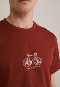 Rūsganas kokvilnas t-krekls ar baltu velosipēda grafiku un tekstu "Kad ceļš tevi aizved" zem tā. Klasiska apaļā kakla dizains.