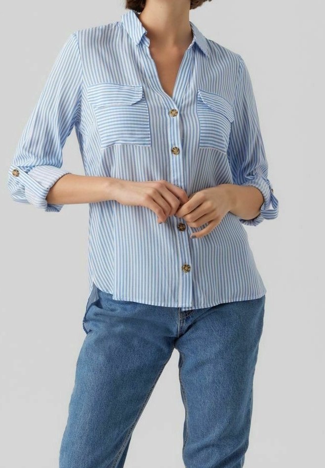 Femme portant une chemise à rayures verticales bleues et blanches avec des manches retroussées et un jean bleu, les mains jointes à la taille.