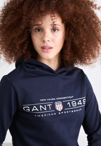 Hanorac bleumarin dintr-un material moale, cu un grafic frontal ce include textul "GANT 1949" și "American Sportswear" în alb, plus un design cu un scut.