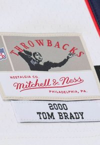 Weißes Stoffetikett mit einer Grafik eines Spielers in Aktion, rotem Text "THROWBACKS" und dem Logo "Mitchell & Ness", das die Jahreszahl "2000" und "TOM BRADY" zeigt.