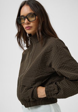 Femme portant de grandes lunettes de soleil carrées et une veste zippée marron à carreaux avec des poignets élastiques et des poches, posant sur fond blanc.
