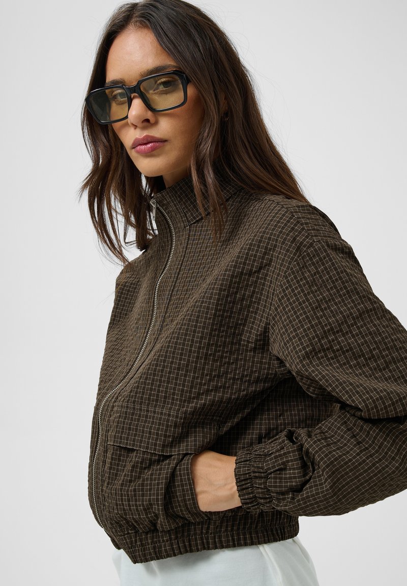 Femme portant de grandes lunettes de soleil carrées et une veste zippée marron à carreaux avec des poignets élastiques et des poches, posant sur fond blanc.