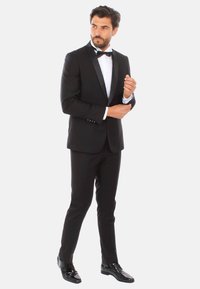 Tuxedo nero con rever in raso, camicia bianca e cravatta a farfalla. Taglio su misura con scarpe eleganti lucide, in posa mentre aggiusta il polsino con la mano destra.