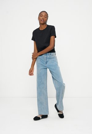 Femme debout devant un fond blanc portant une chemise noire à manches courtes, un jean large bleu clair et des ballerines noires avec des brides blanches.