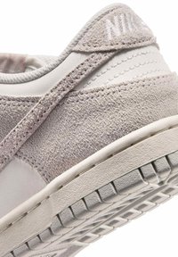 Nærbillede af en beige og hvid sneakers hæl, der viser struktureret ruskind, syede læderpaneler og en mønstret gummisål.