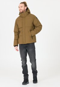 Brun pufferjakke med lynlås foran, høj krave og sidelommer. Båret med grå jeans og sorte vandrestøvler.