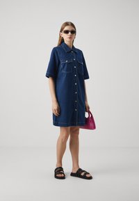 Robe chemise en denim bleu foncé, à manches courtes, avec deux poches poitrine et des boutons à l'avant. Associée à des sandales noires et tenant un sac rose.