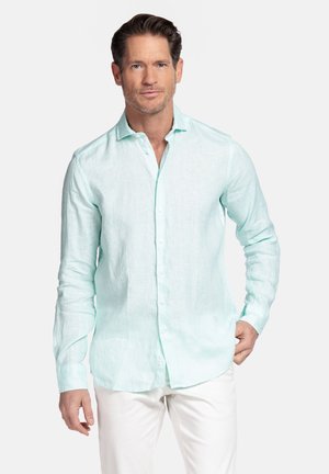 Man met lichtblauw overhemd met lange mouwen en witte broek, staand tegen een effen lichte achtergrond.