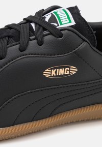 Svarta lädersneakers med en sula av gummigranulat, stickade detaljer och en metallisk guld "KING"-logotyp på sidan. Grön och vit märkesetikett ovanför.