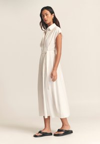 Witte jurk met kraag, korte mouwen, een taille met strik en een vloeiende silhouet. Draagt zwarte platte sandalen. Glad oppervlak.