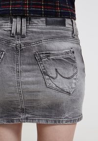 Šedá džínová sukně s poškozenou texturou, vybavená dvěma zadními kapsami a jemným zakřiveným švem. Značka viditelná na pásku.