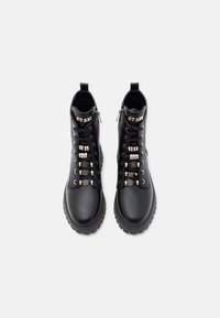 Bottes de cheville en simili cuir noir avec des œillets métalliques décoratifs, semelle en caoutchouc épaisse et une fermeture éclair de chaque côté. Logo "GUESS" sur la languette.