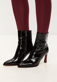 Zwarte lakleren enkellaarsjes met een spitse neus en een slim, rood hakje. Voorzien van een zijrits en een nauwsluitende pasvorm, met een glanzende, gladde textuur.