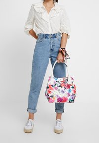 Květinová kabelka s bílým podkladem, která má živé vícobarvené květinové vzory, zaoblené uchy a strukturovaný tvar.
