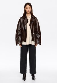 Mujer con una chaqueta de cuero de gran tamaño marrón oscuro, camisa beige con botones, pantalones negros holgados y botas negras de punta, de pie contra un fondo blanco.