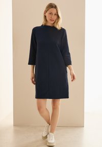 Marineblaues Tunika-Kleid aus strukturiertem Stoff mit Dreiviertelärmeln, einem runden Ausschnitt und gerader Passform, kombiniert mit weißen Turnschuhen.