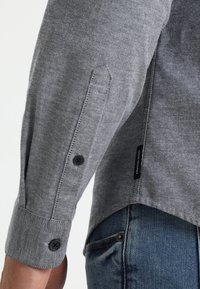 Szara tkanina koszulowa z zapinanymi na guziki mankietami, z subtelną fakturą i zaokrąglonym dołem. Widoczna metka marki w pobliżu dolnego szwu.