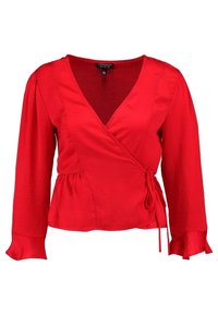 Blouse portefeuille rouge à manches longues, encolure en V et fermeture à nouer. Confectionnée dans un tissu lisse ; manches ornées de volants au niveau des poignets.