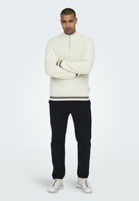 Sudadera blanca con cremallera, textura acanalada y acento de rayas negras en el dobladillo, combinada con pantalones negros y zapatillas blancas.