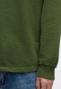 Πράσινο μακρυμάνικο πουκάμισο με υφή υφάσματος, ριμπ μανσέτες και διπλή διακόσμηση με κυματιστό τελείωμα στο κάτω μέρος. Συνδυάζεται με μπλε τζιν παντελόνι.