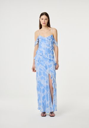 Vrouw draagt een lichtblauwe, off-shoulder, gerimpelde maxi-jurk met een dijhoge split en dunne bandjes, staand tegen een effen achtergrond.