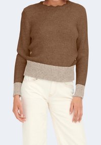 Brauner gestrickter Pullover mit geripptem beigem Saum und Bündchen, ausgestattet mit einem gewellten Ausschnitt und strukturiertem Muster. Kombiniert mit hellen Jeans.