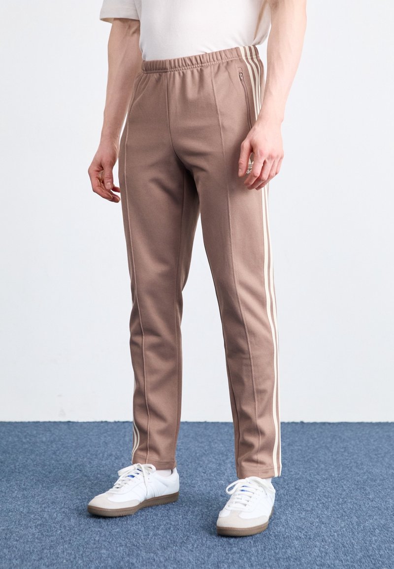 Pantaloni sportivi marroni con strisce laterali bianche, vita elastica, taglio affusolato e texture morbida, abbinati a sneakers bianche.